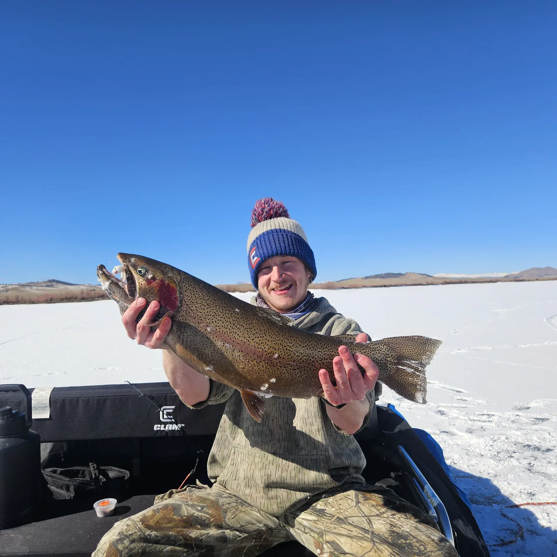 An angler seated on a gear sled holding an outsized trout under a clear blue sky