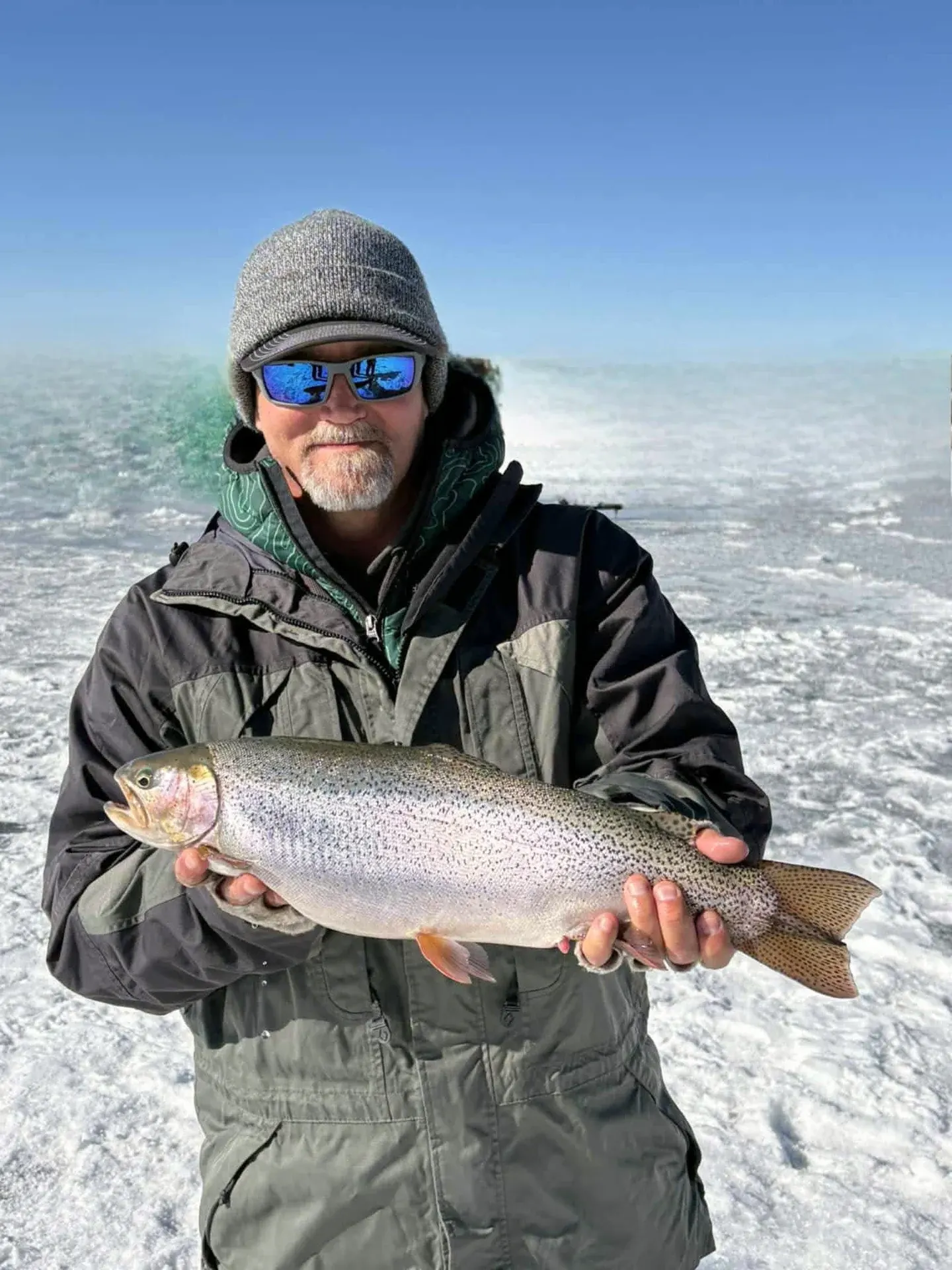 An angler on the ice holding a large rainbow trout, the frozen reservoir behind him