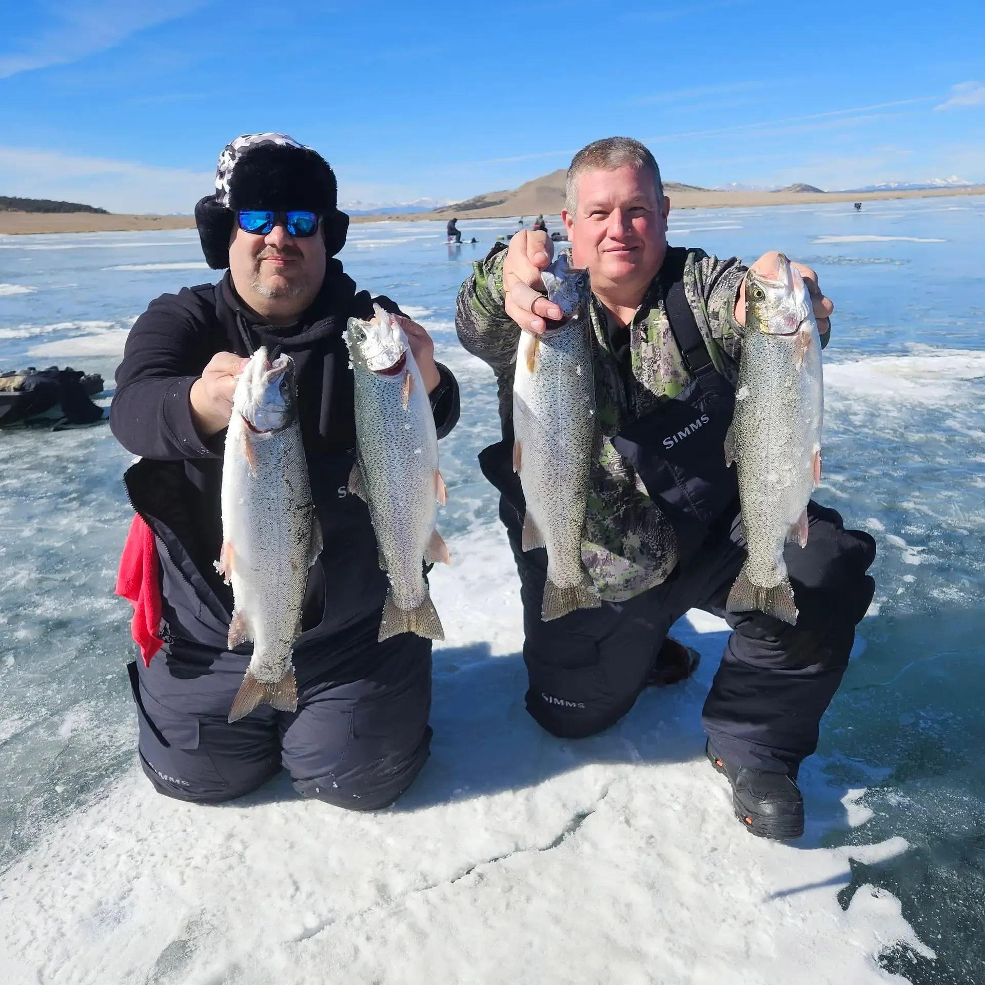 Two anglers kneeling on the frozen reservoir, each holding a pair of trout